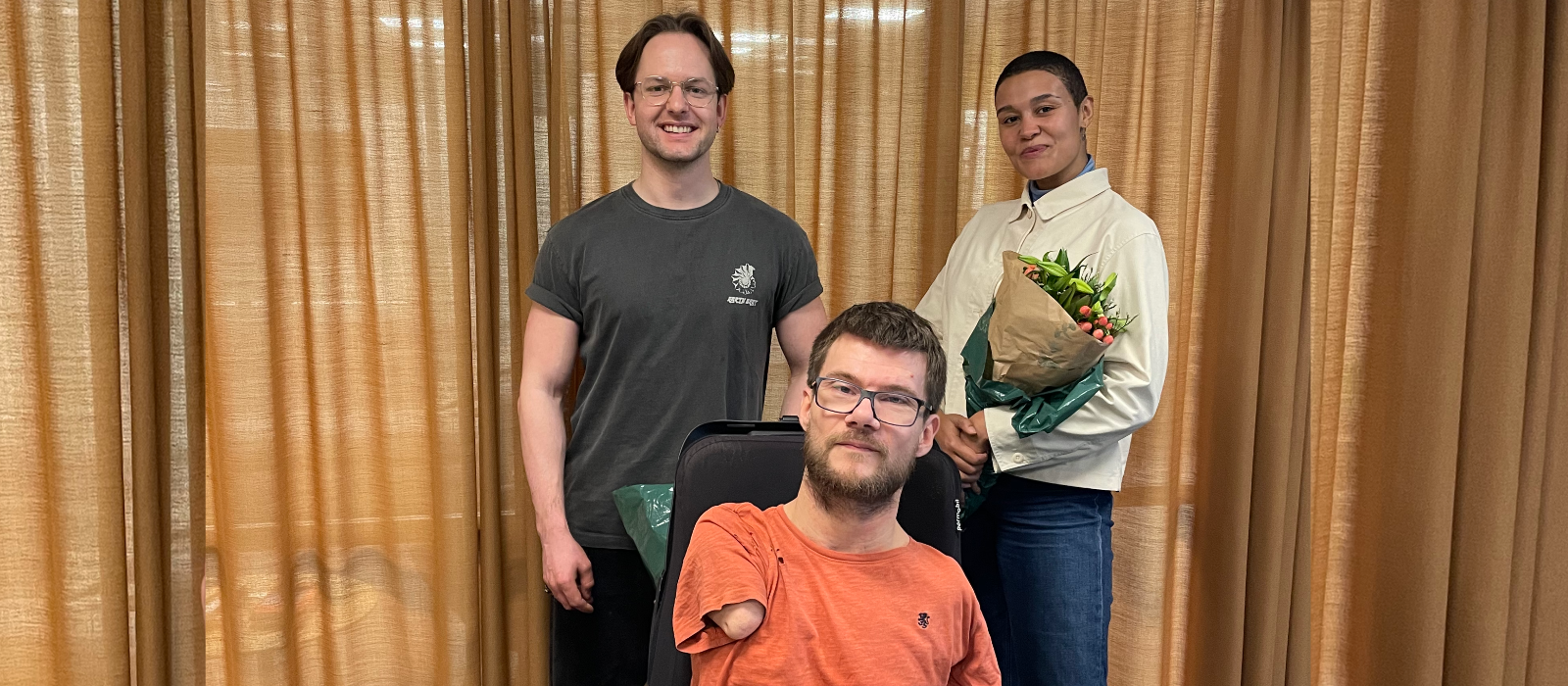 Tre av Arrangørprisens jurymedlemmer poserer etter årets jurymøte. Personen i front sitter i en elektrisk rullestol og personen bakerst til høyre holder en blomsterbukett. 