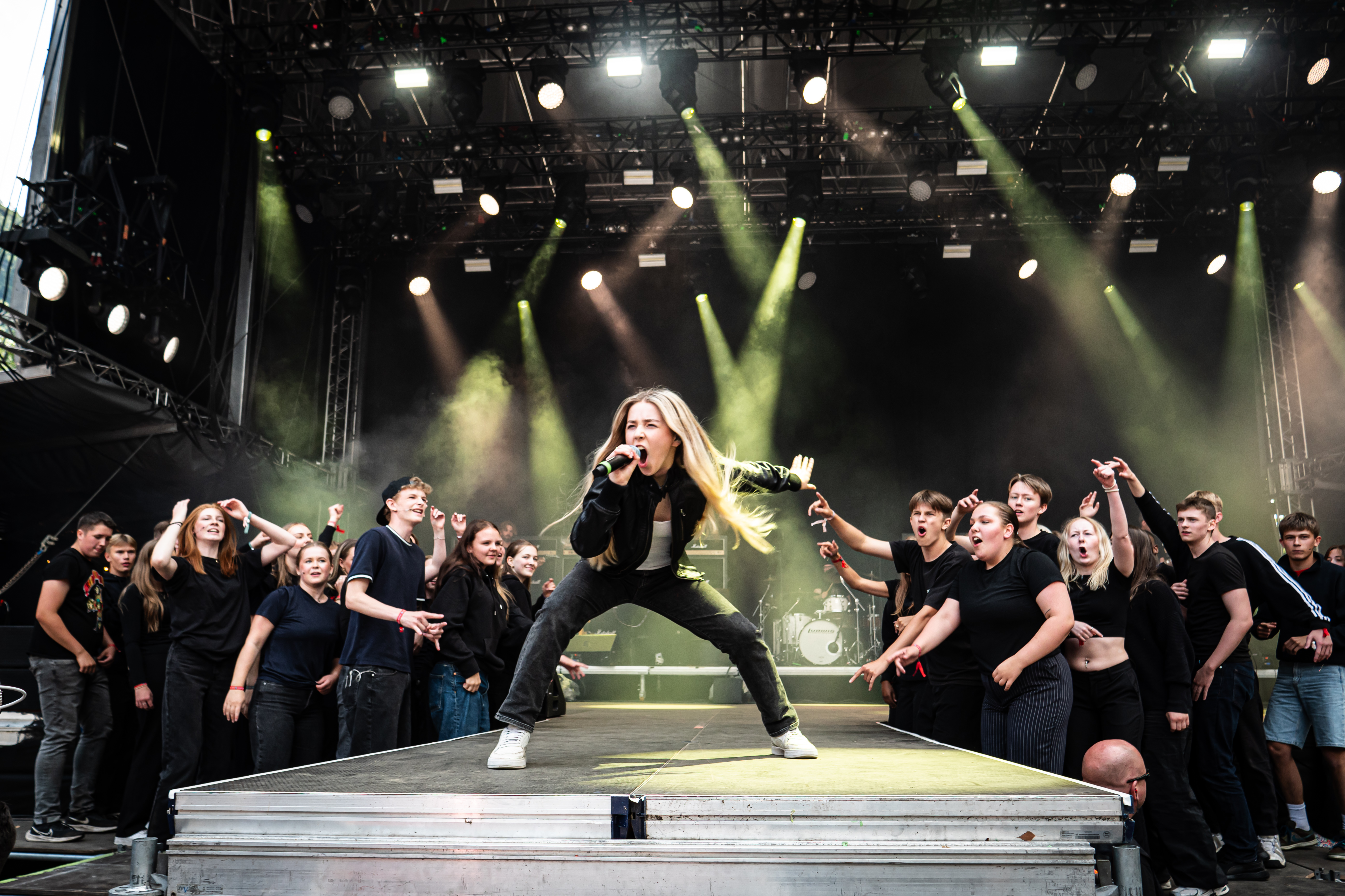 Eksplosiv fremføring av en stor gruppe ungdommer i helsvarte antrekk. I front en kvinnelig vokalist som synger med full kraft. I bakgrunnen, på hver side av scenen er det en større gruppe ungdommer som synger og danser med.