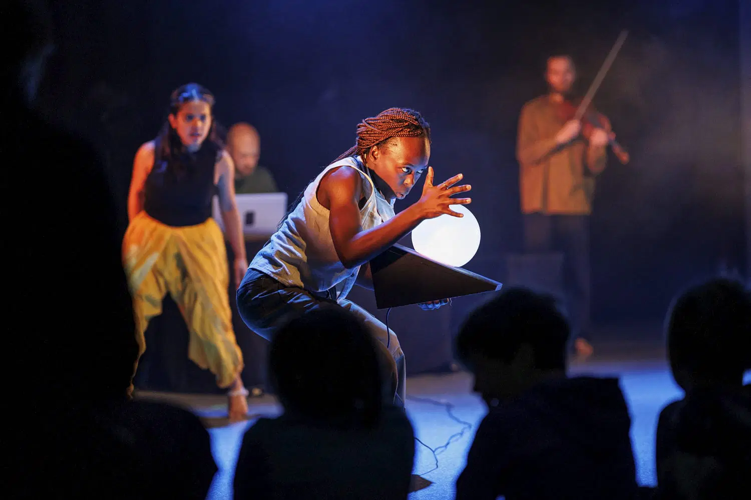 En danser beveger seg med intenst blikk over scenen. I bakgrunnen står musikere og en annen danser. Siluetter av barn i publikum fyller forgrunnen. 