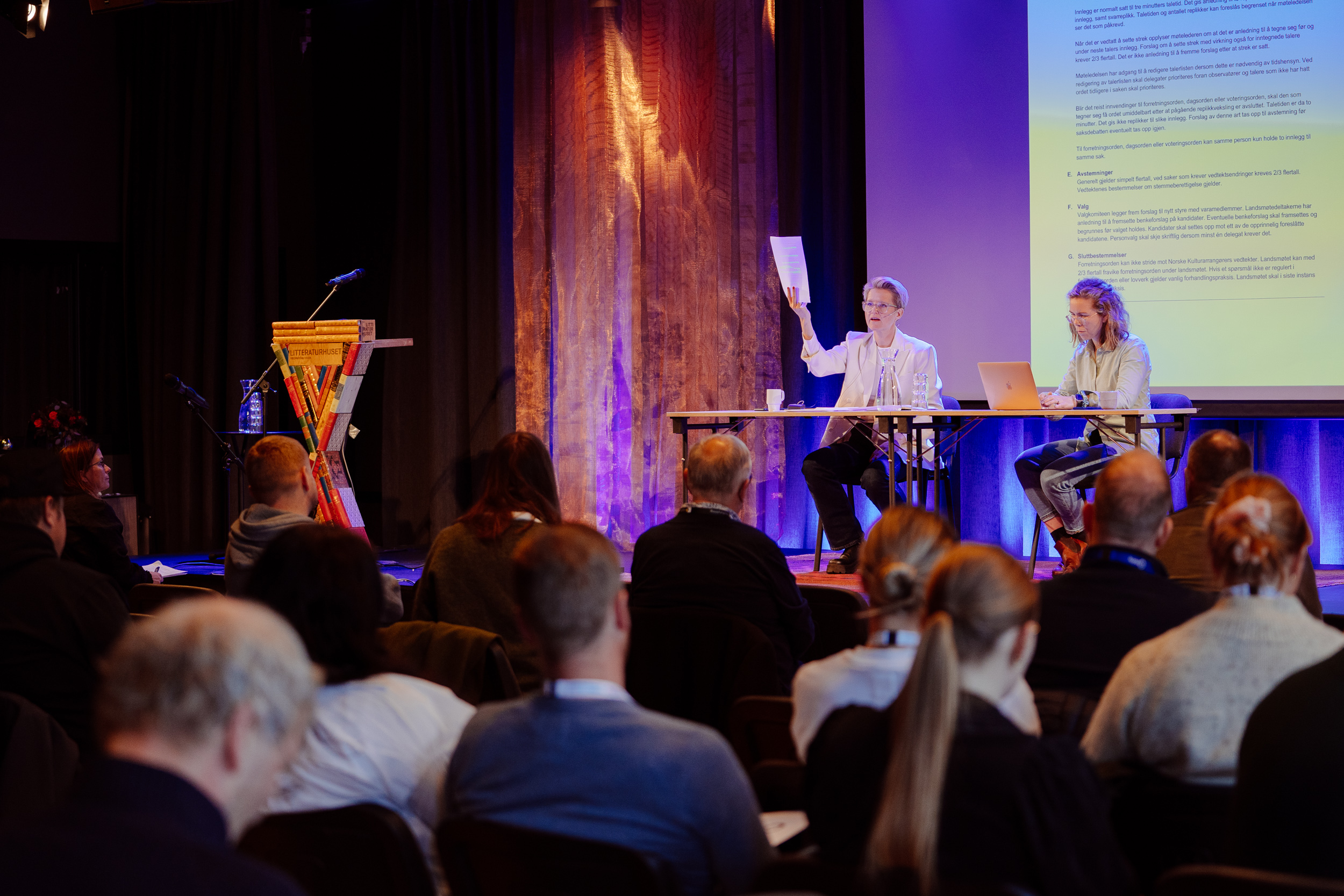 Ordstyrer og bisitter forklarer saksgang under Norske Kulturarrangørers landsmøte i 2024. I salen sitter medlemmer og lytter. 