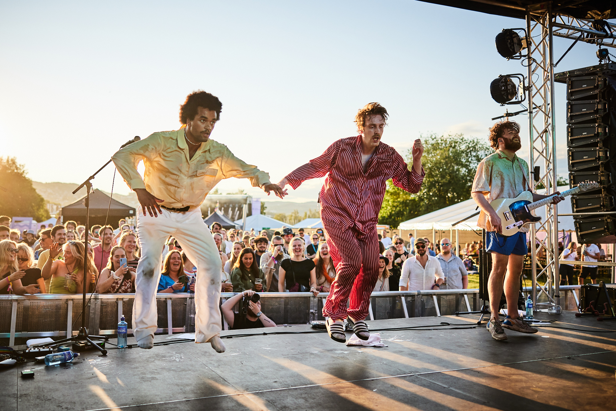 Tre personer hopper energisk med ryggen mot publikum på en festivalscene. Kveldssol og ivrige publikummere i bakgrunnen. 
