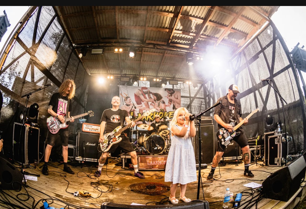 På scenen står et punkband bestående av fire svartkledde voksne menn. På vokal i front står en liten jente i hvit kjole og synger. 