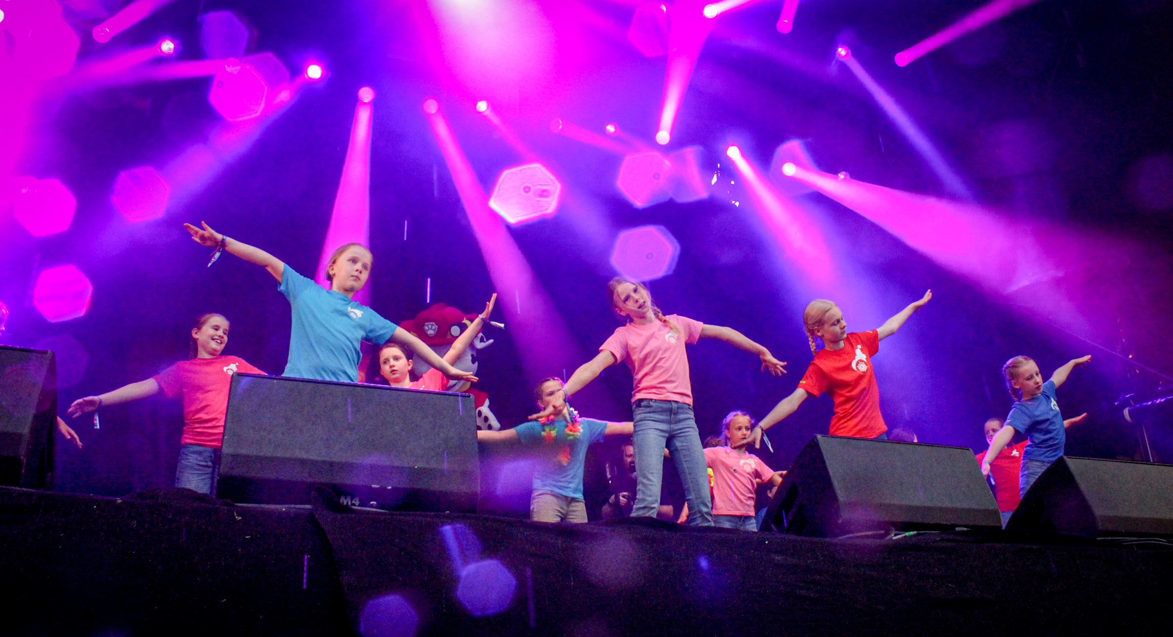 Bildet viser et arrangement på Kulturbanken. Ni barn står på en scene og fremfører en dans. De har på seg t-skjorter i ulike farger: turkis, rosa, rød og blå. De står med armene strakt ute i formasjon.  