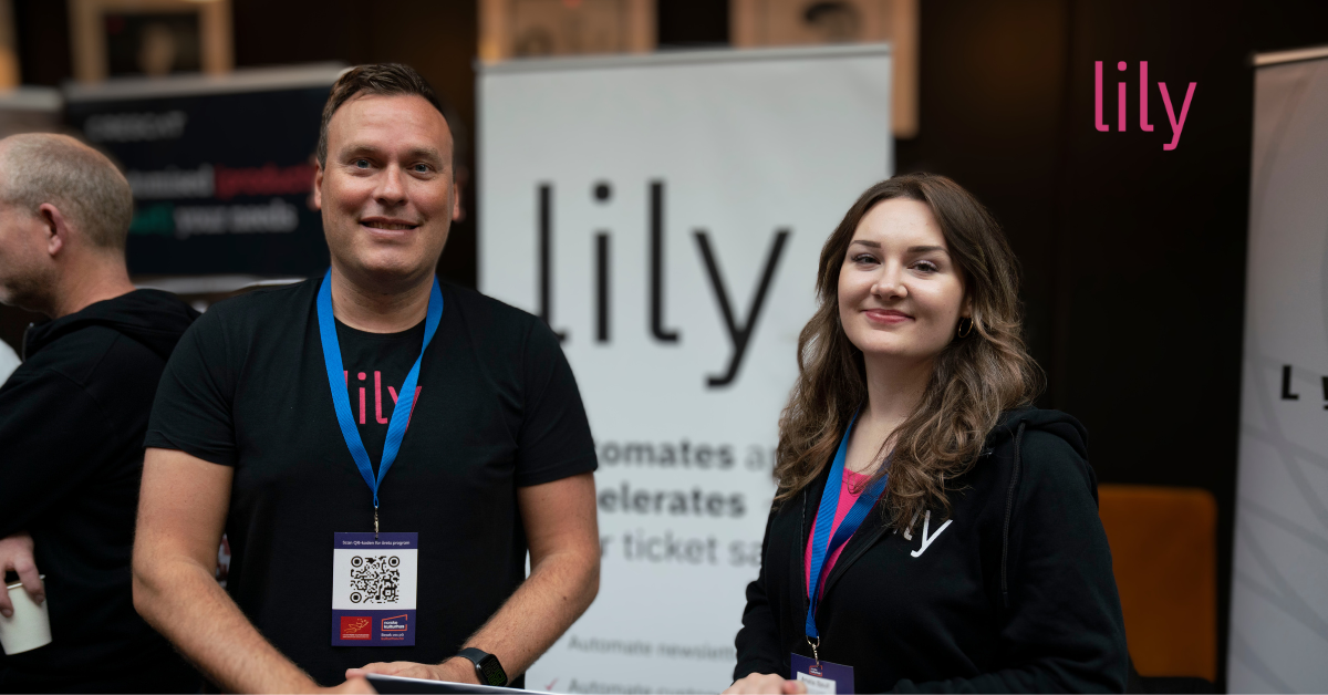 To ansatte fra Lily står på stand og smiler. De er ikledd klær med Lilys logo. I bakgrunnen ser vi rollups med logo og infomasjon om Lilys tjenester. 