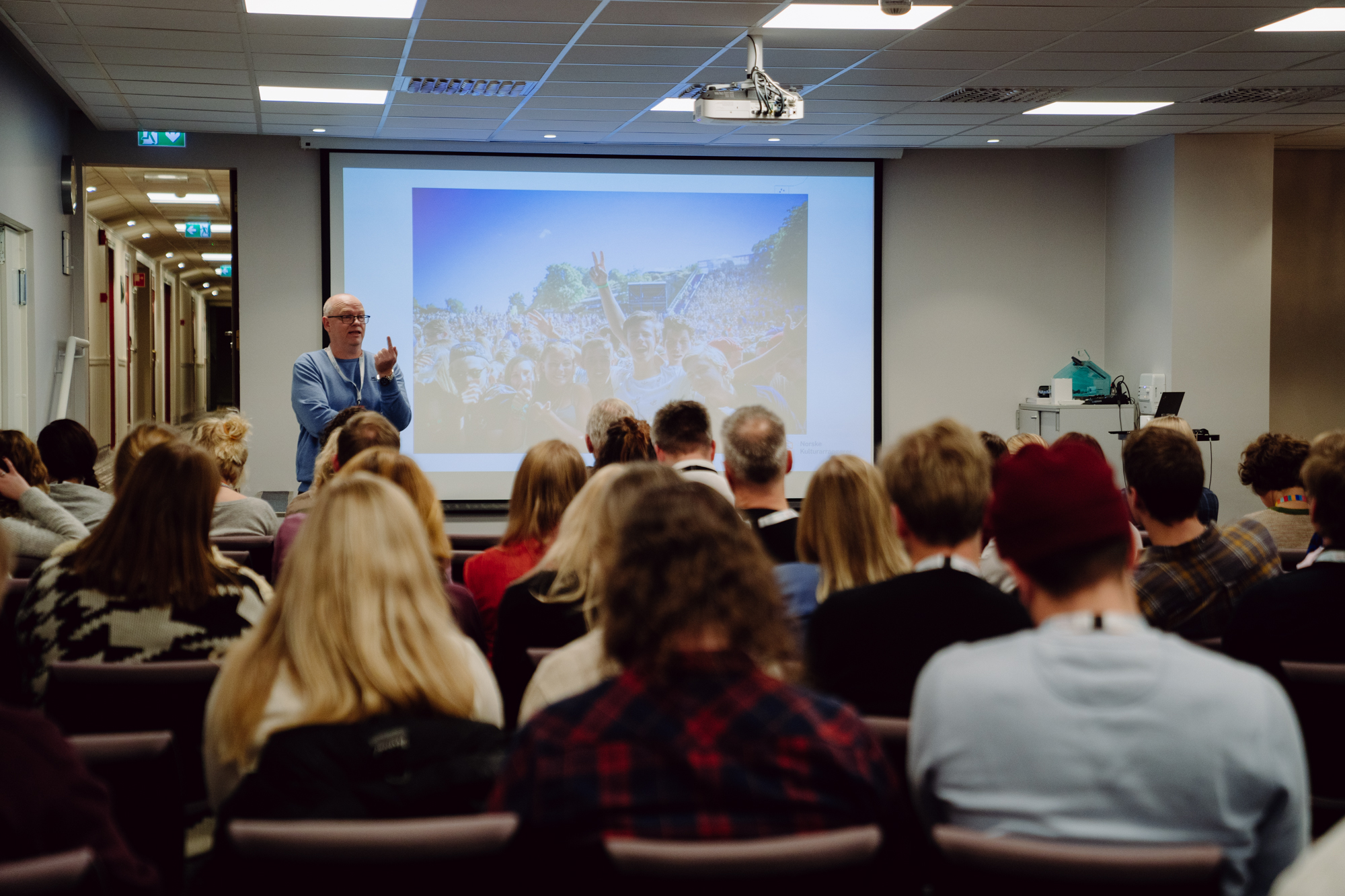 En engasjert kursholder fra Norske Kulturarrangører holder kurs foran en gruppe som sitter plassert ut i kinooppsett i et opplyst møterom. I bakgrunnen skimtes en presentasjon med bilde av glade festivalgjengere. 