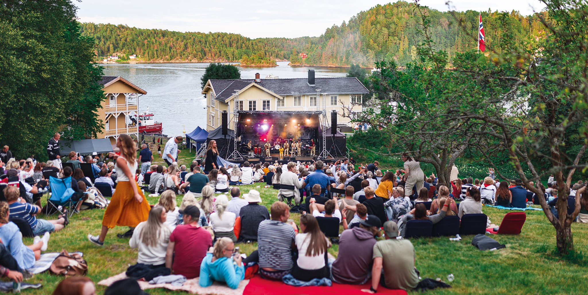 I idylliske og grønne omgivelser ved vannet sitter publikum spredt på pledd og campingstoler utover en gresslette og nyter en konsert en sommerkveld. I bakgrunnen skimtes gamle ærverdige trehus. 