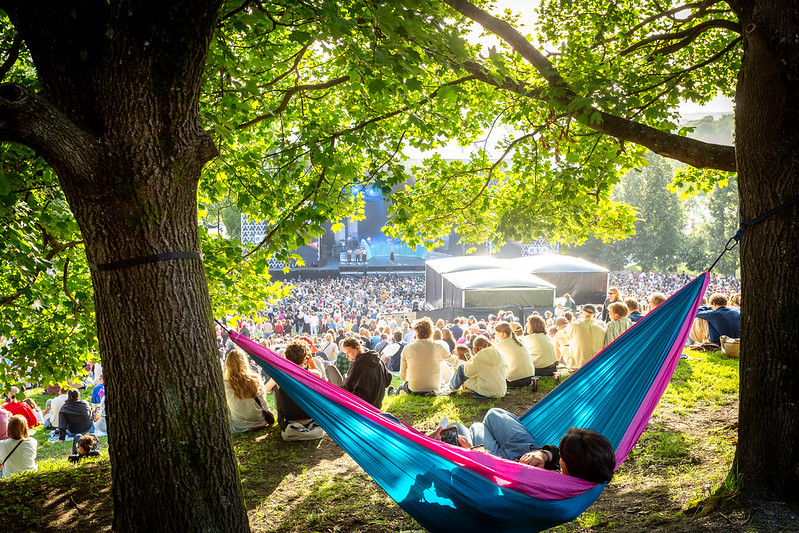 Sol og sommerstemning på Øyafestivalen. Mellom to trær ligger to personer i en hengekøye og slapper av mens vi skimter hovedscenen og et folkehav som nyter festivalen i bakgrunnen. 