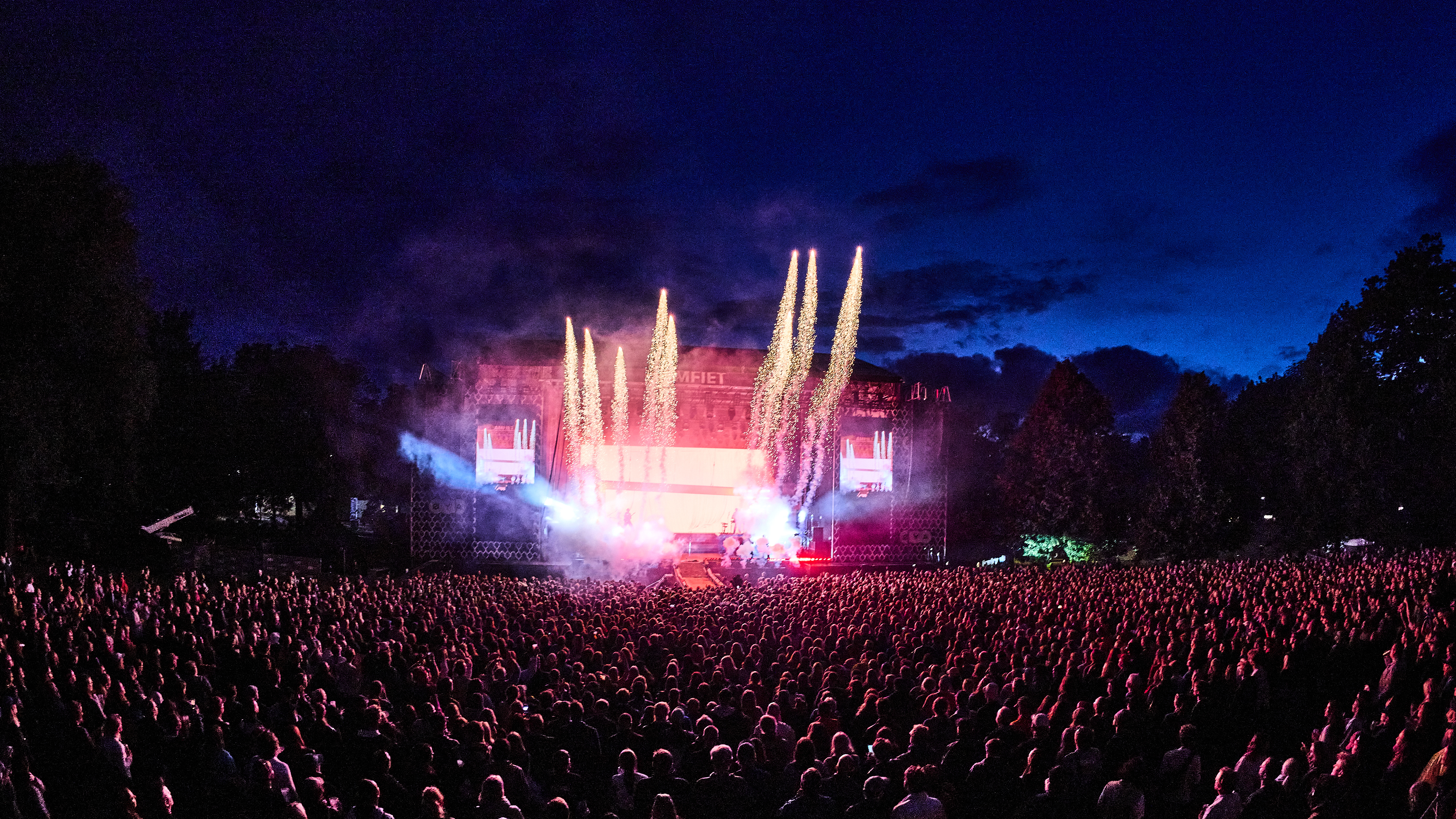 Oversiktsbilde av en folkemengde foran en utendørs scene med lysshow og fyrverkeri.