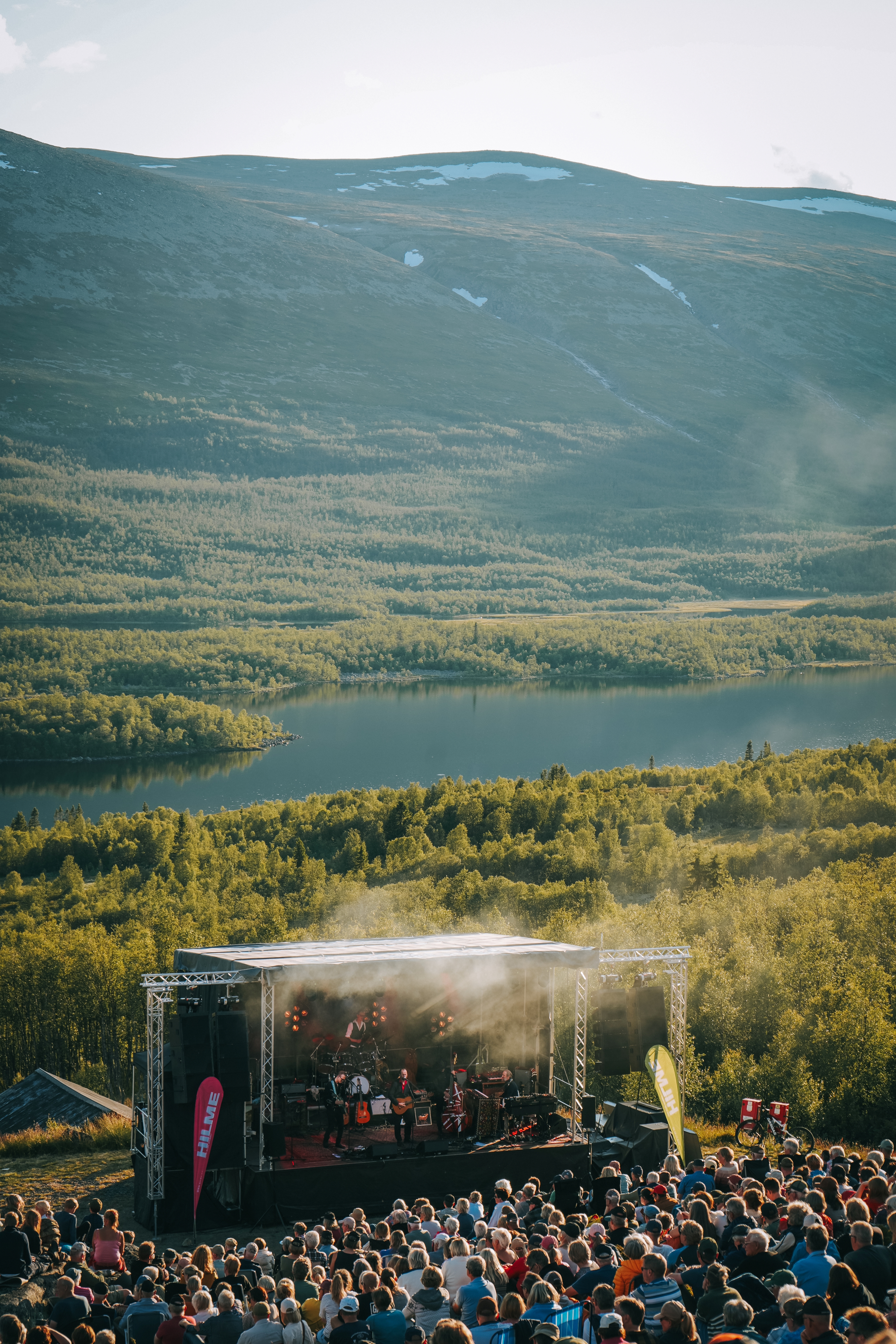 Oversiktsbilde av en utendørs konsertscene i grønne omgivelser med en folkemengde foran.